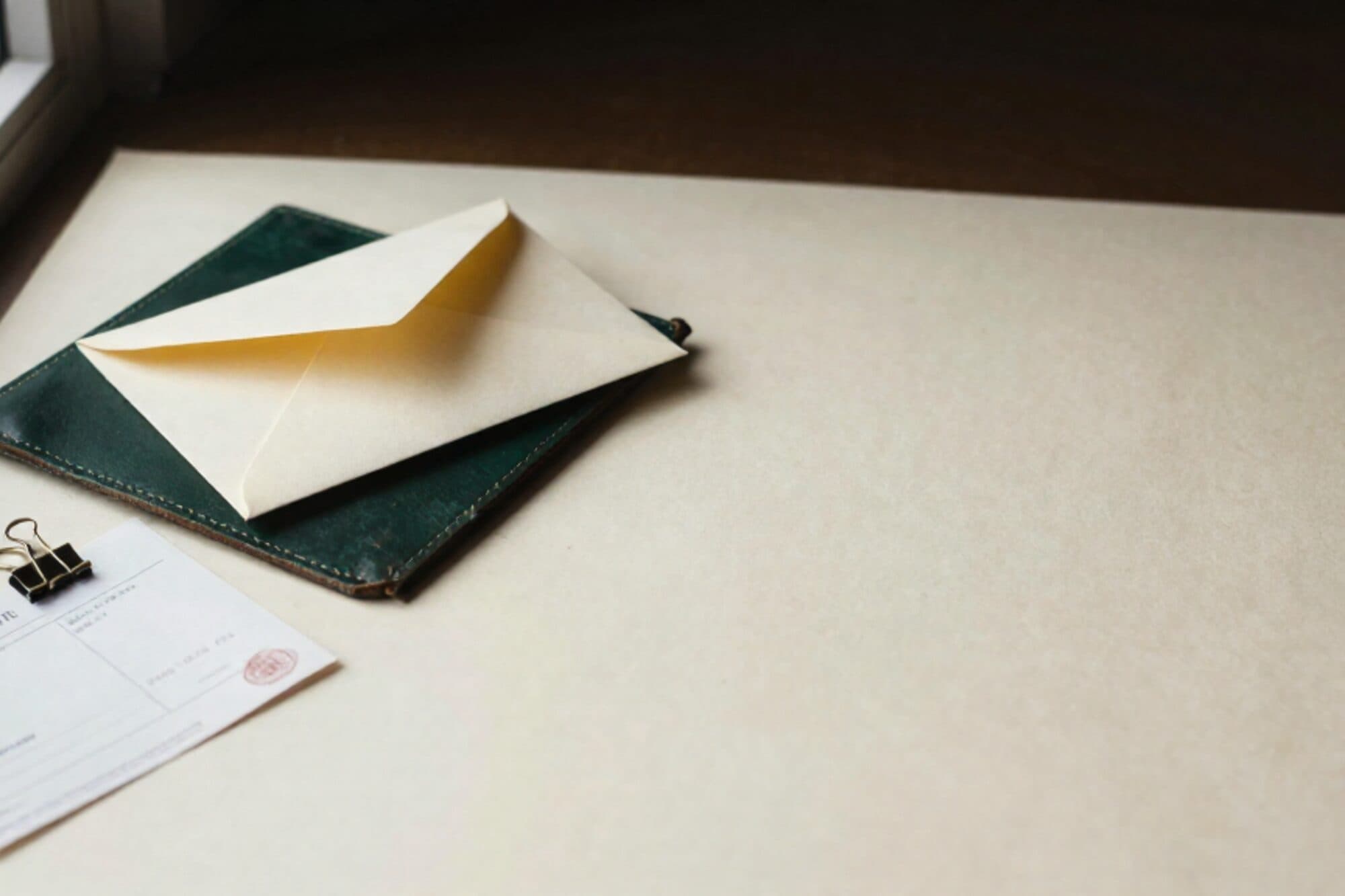 Crème envelop met limegroen lint op donkergroen leren onderlegger, koperen paperclip, gevouwen doorslagfactuur ernaast.